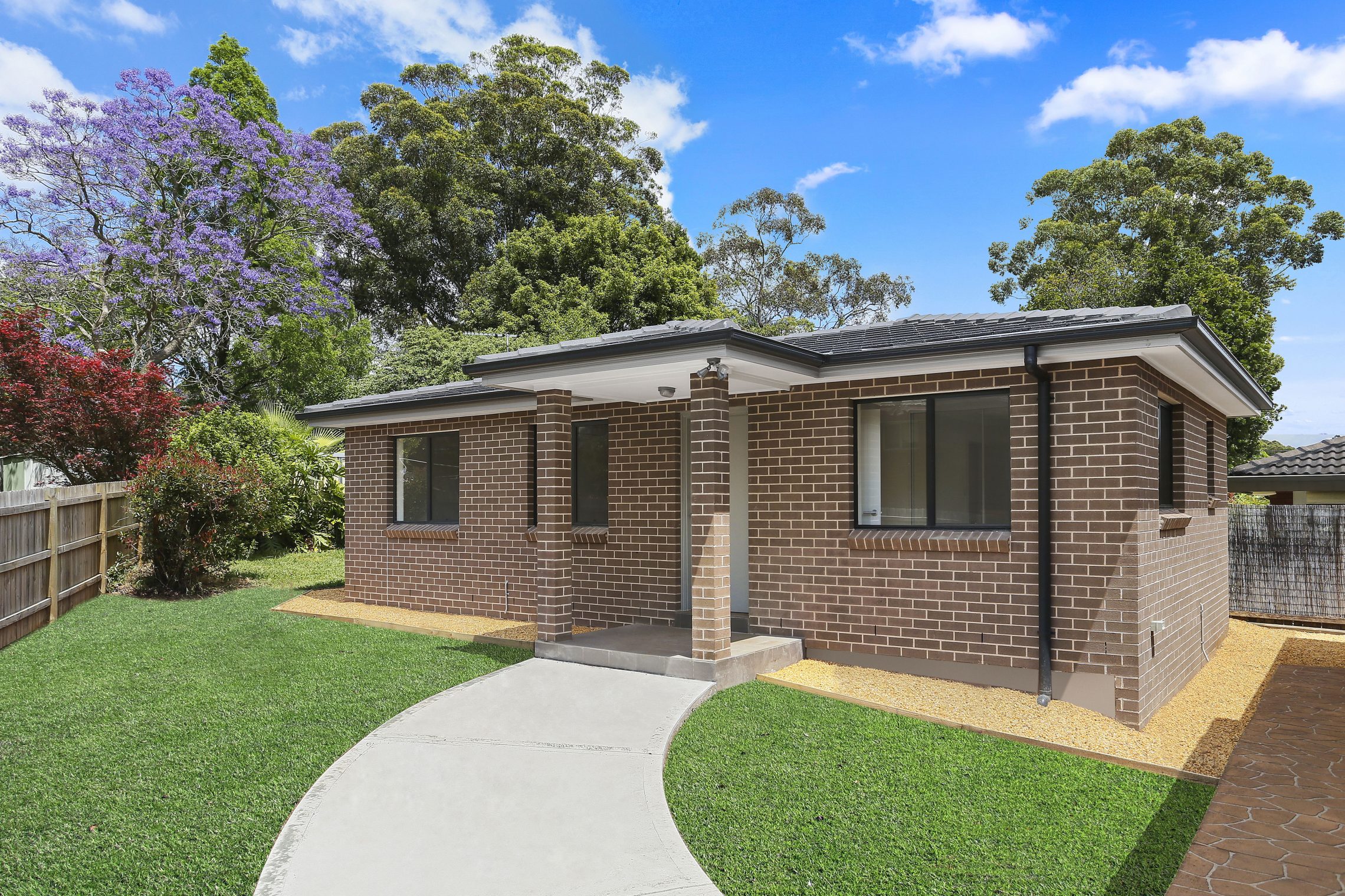 Dark brick granny flat, surrounded by trees, with cement walkway leading to covered entry